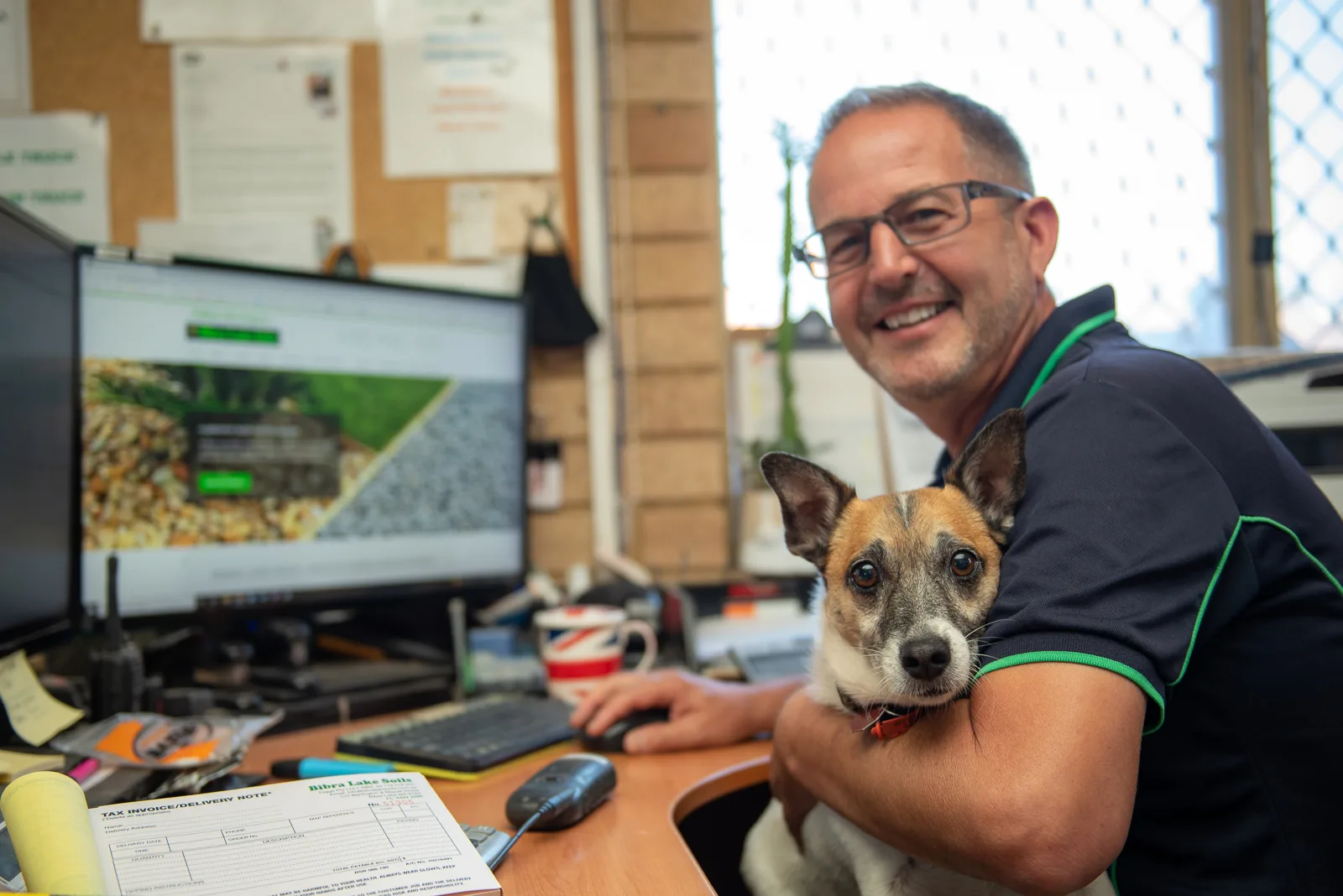 A Bibra Lake Soils staff member at the office desk taking an order, with one of the yard dogs on his lap